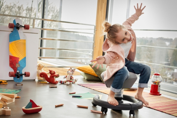 Mädchen balanciert barfuß mit ausgestreckten Armen auf einem beweglichen Sitz in einem Kinderzimmer.